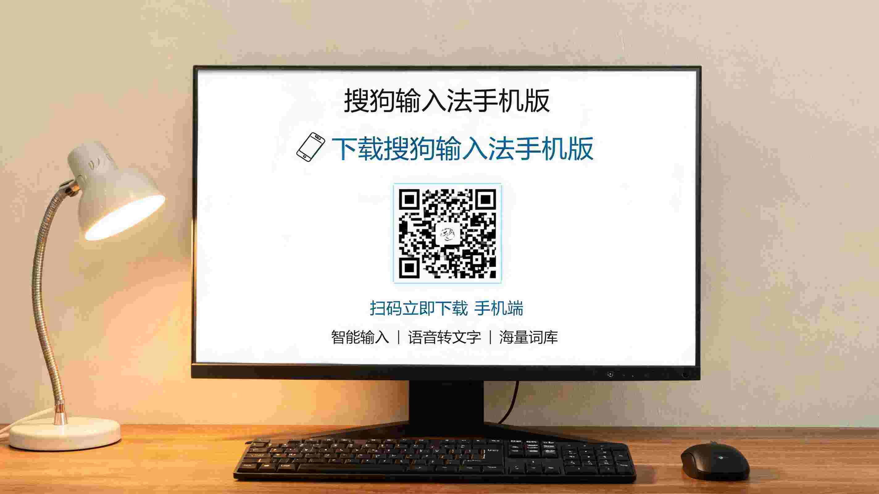 电脑桌上的电脑，屏幕显示搜狗输入法手机版下载界面，旁边放着台灯